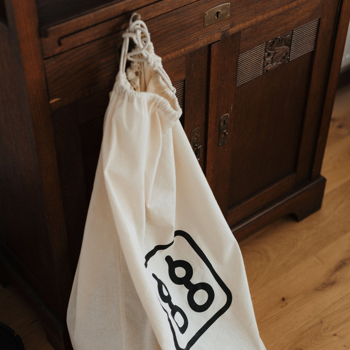 Cremefarbener Aufbewahrungsbeutel mit schwarzem Logo-Aufdruck, an dunklem Holzschrank hängend, Parkettboden.