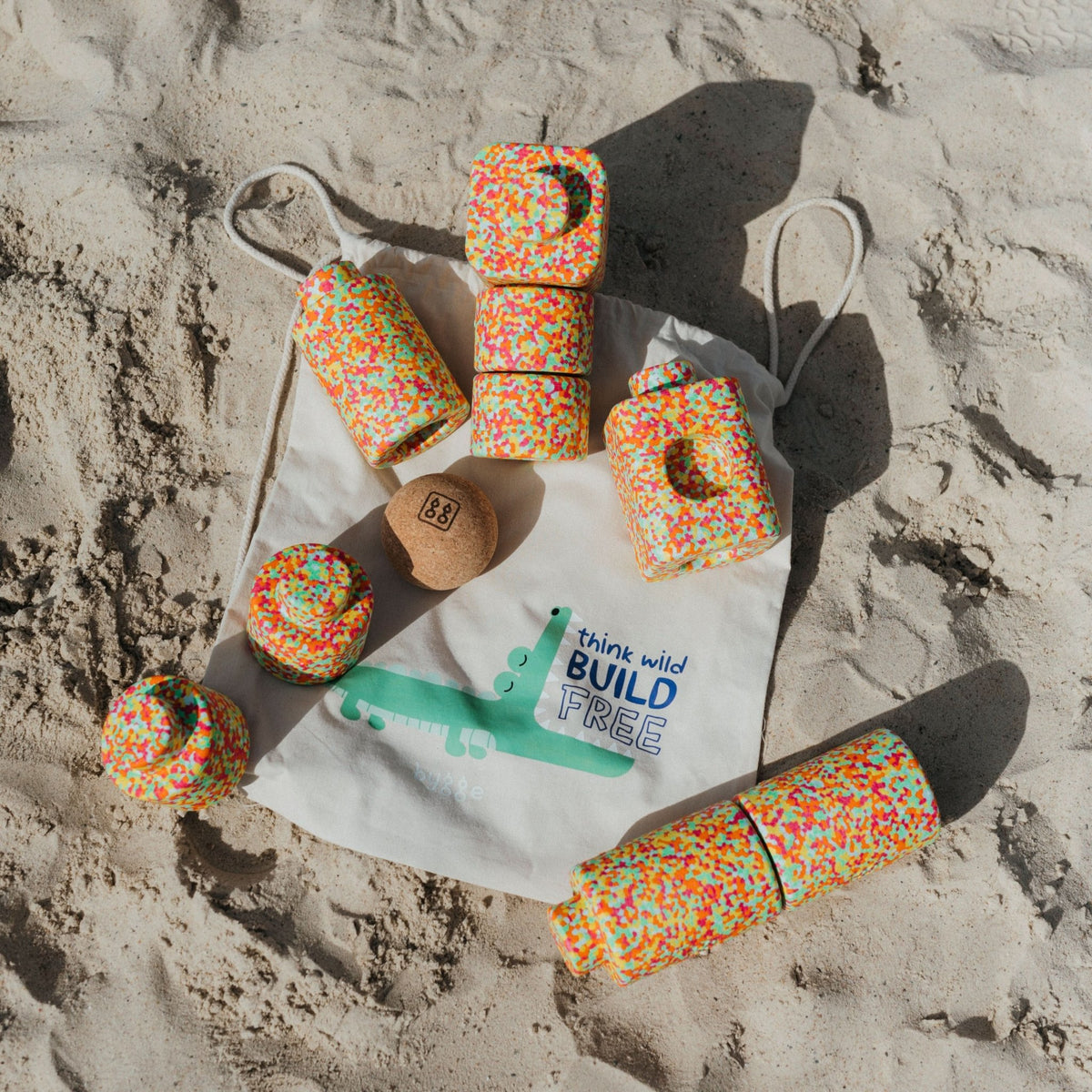 Bunte EPP-Bausteine XXL in Konfetti-Optik auf Sand, daneben Stoffbeutel mit Krokodil-Motiv und Korkball.