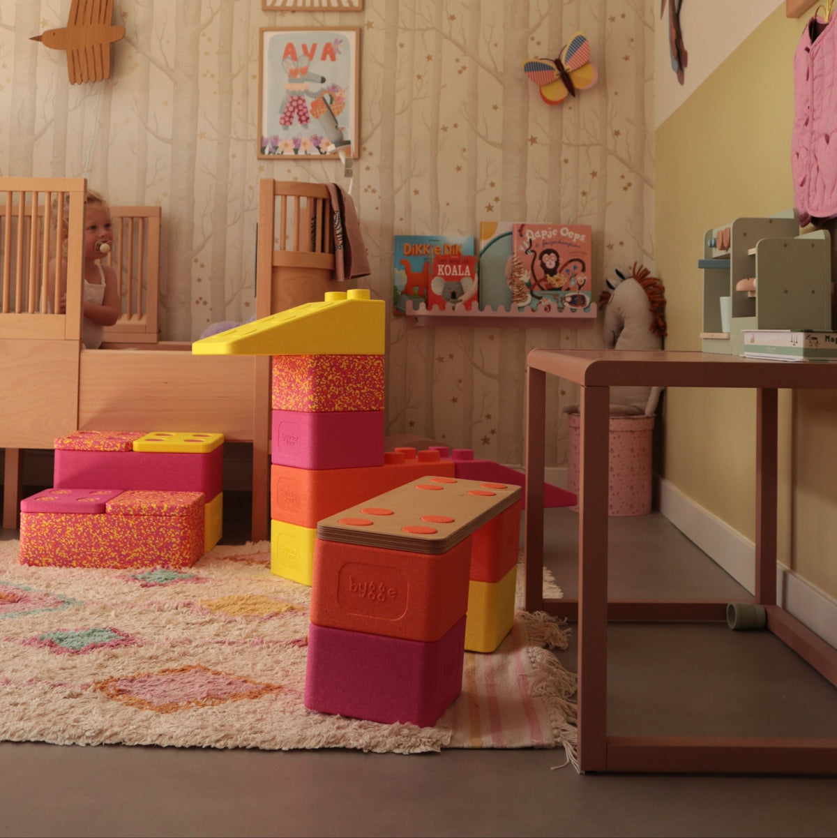 Kleinkind im Bett beobachtet bunte bygge XXL-Bausteine in Pink, Orange, Gelb – als Turm und Treppe aufgebaut im Kinderzimmer.