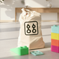 Cremefarbener Aufbewahrungsbeutel TASKE mit Logo, daneben bunte XXL-Bausteine in Grün, Blau, Gelb, Pink und Orange.