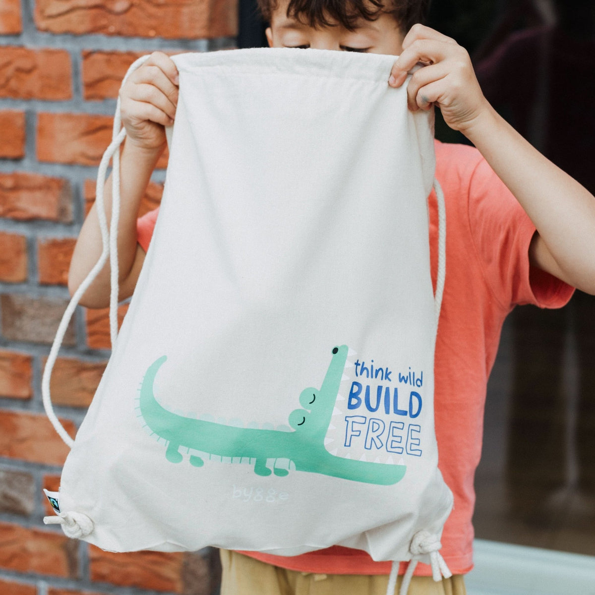 Kind hält weißen Beutel "KROKO" mit türkisem Krokodil-Aufdruck und Schriftzug "think wild BUILD FREE" hoch.