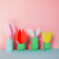 4 bunte Papierblumen in mintgrünen und grünen Schaumstoffvasen vor rosa Hintergrund: rosa, rot, weiß und gelb.