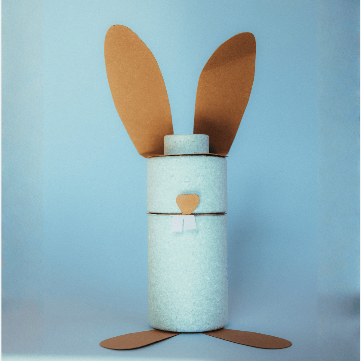 Mintgrüne EPP-Baustein-Zylinder als Osterhase gestaltet: braune Papierohren, Nase und Pfoten auf hellblauem Hintergrund.