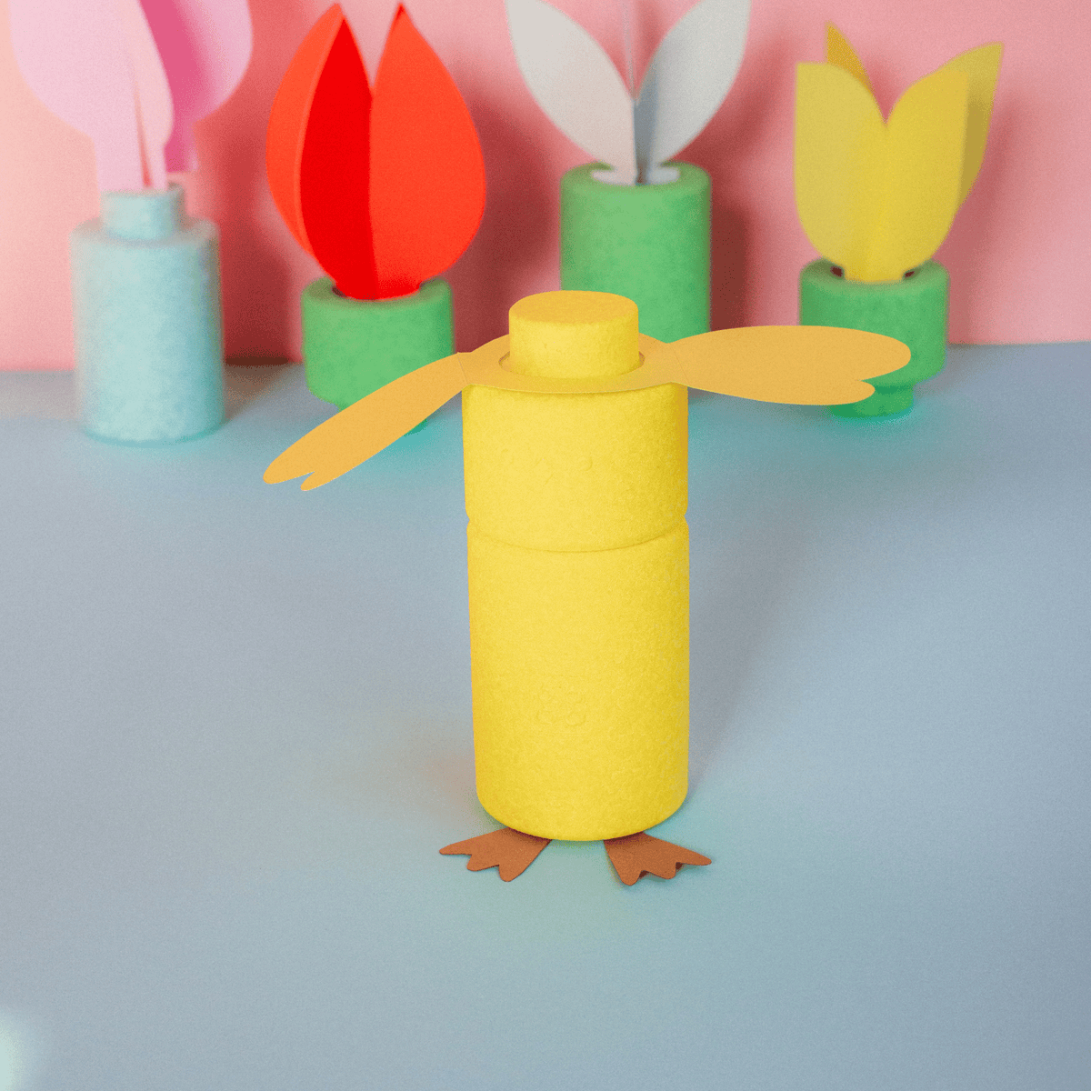 Gelbes Küken aus EPP-Bausteinen mit Papierflügeln und Füßen, dahinter bunte Papierblumen auf blauem Untergrund.