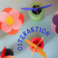 Bunte Papierblumen und EPP-Bausteine XXL auf hellblauem Untergrund mit Schriftzug „Osteraktion