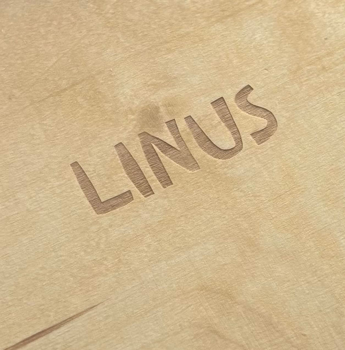 Nahaufnahme eines hellen Holzbretts mit eingefrästem Schriftzug „LINUS" in Großbuchstaben.