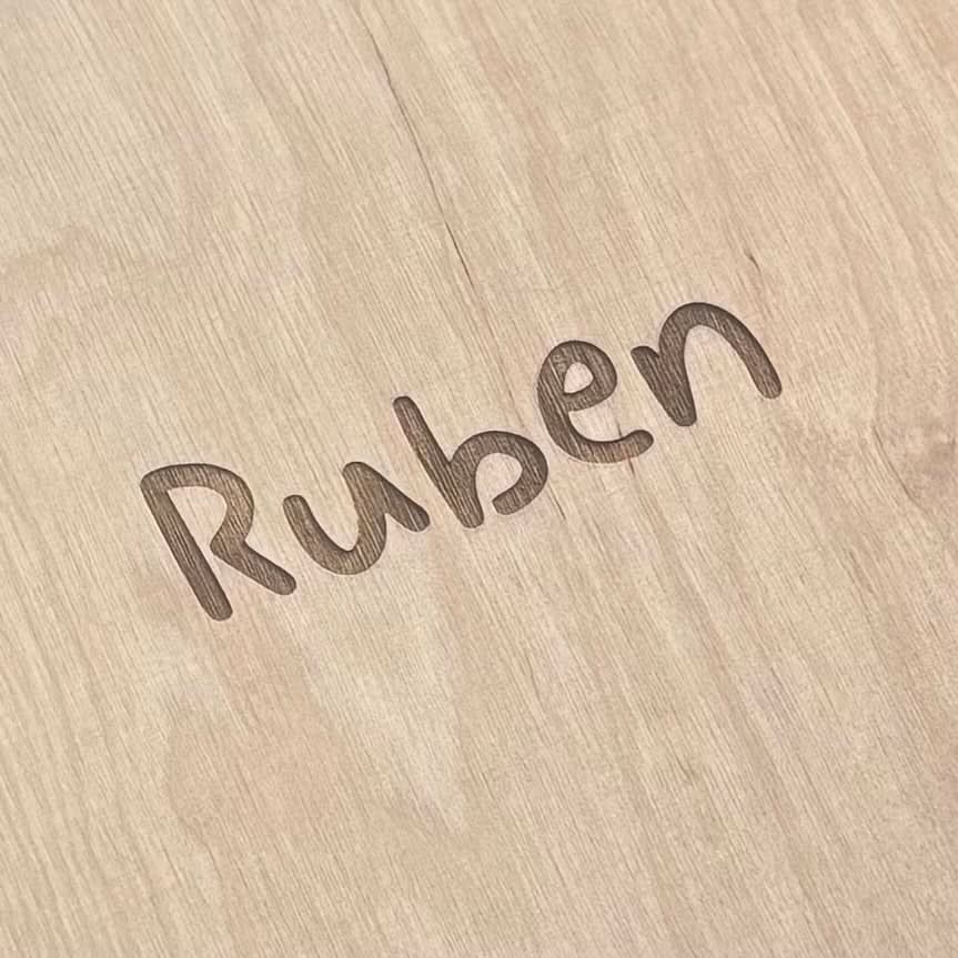 Name „Ruben" in geschwungener Schrift in helles Holz eingraviert – Nahaufnahme einer personalisierten Holzoberfläche.