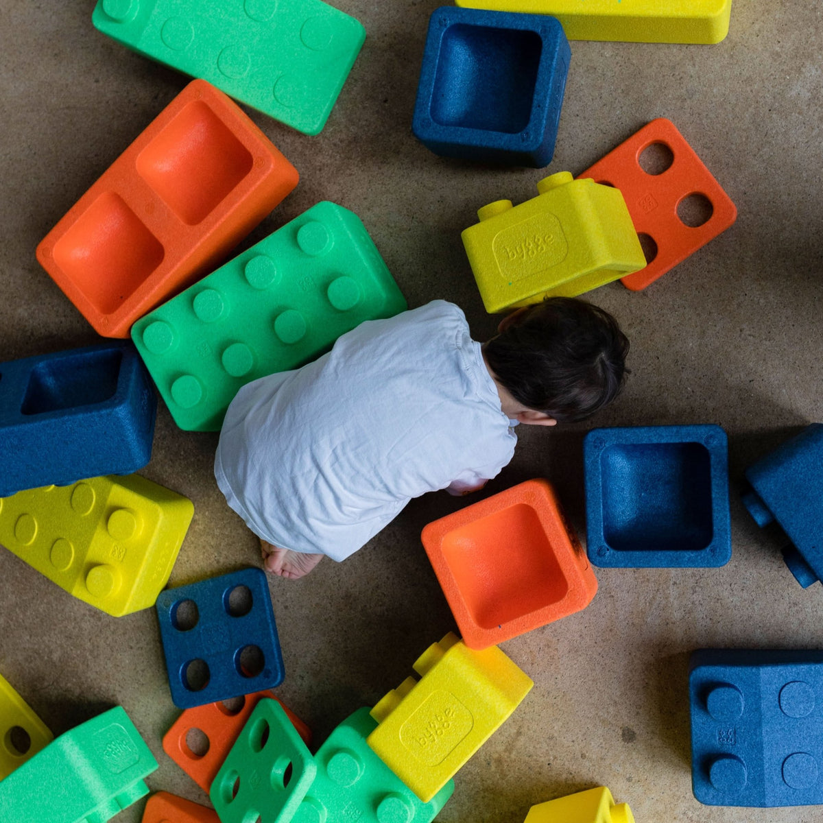 Kleinkind liegt zwischen bunten bygge EPP-Bausteinen XXL in Grün, Blau, Orange und Gelb auf dem Boden.